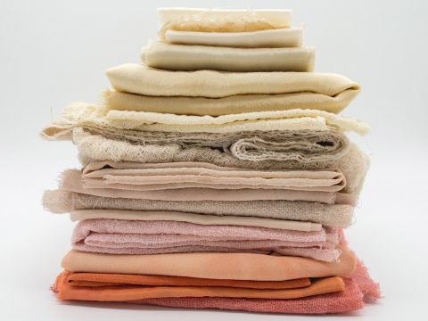 Ein ordentlich gestapelter Stapel verschiedener Stoffe in verschiedenen Creme-, Beige-, Pfirsich- und Orangetönen vor einem schlichten weißen Hintergrund. Die Texturen reichen von glatt bis leicht zerknittert.