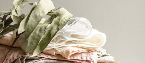 Auf einem Stapel weicher, pastellfarbener Stoffe liegt ein großes, getrocknetes grünes Blatt. Der Hintergrund ist in neutralem Beige gehalten und hebt die Texturen und Farben der Stoffe und des Blattes hervor.