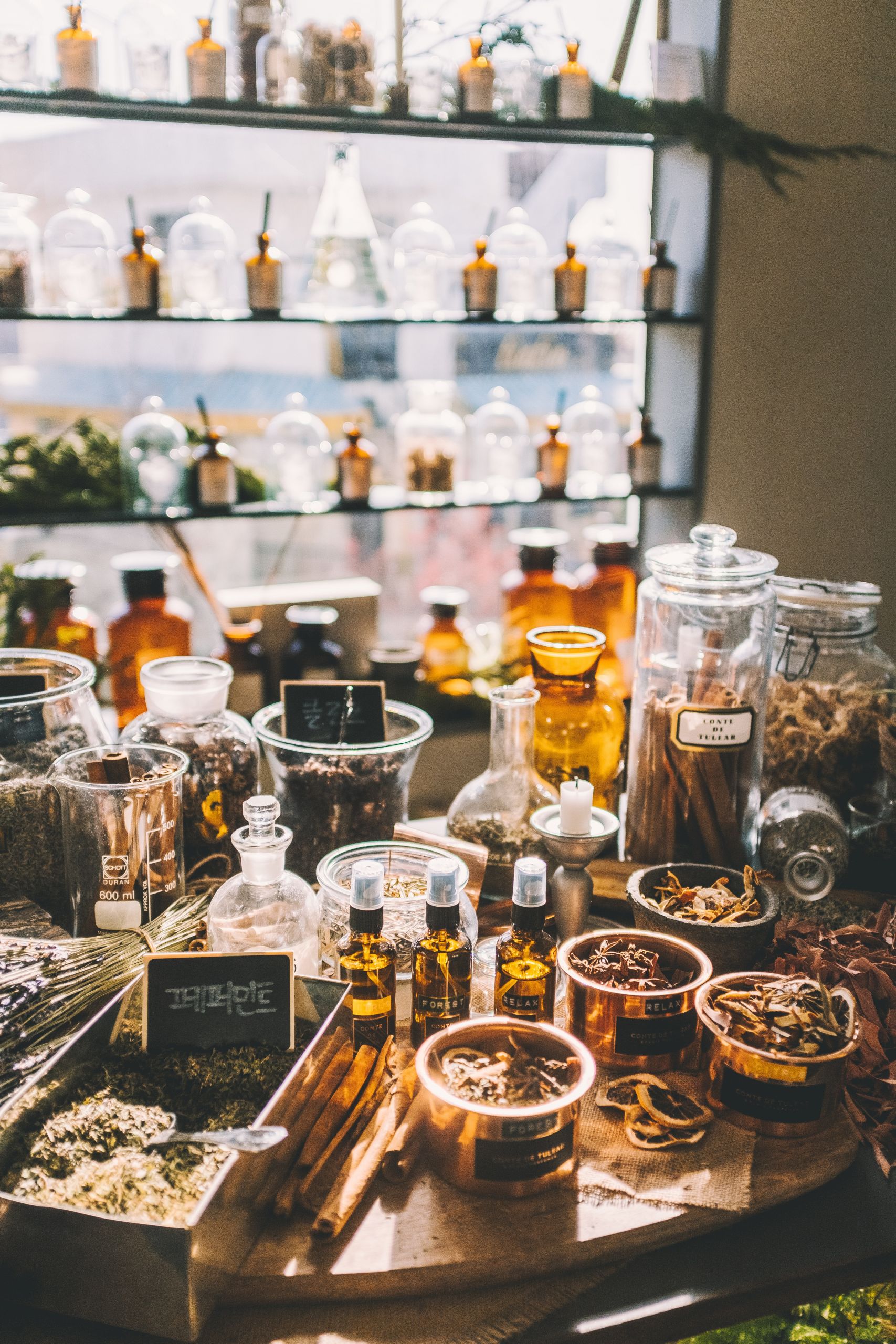 Eine lebendige Apothekenauslage mit verschiedenen Gläsern und Flaschen, gefüllt mit Kräutern, Gewürzen und ätherischen Ölen. Im Hintergrund sind Regale mit weiteren Flaschen zu sehen, die eine warme, aromatische Atmosphäre schaffen.