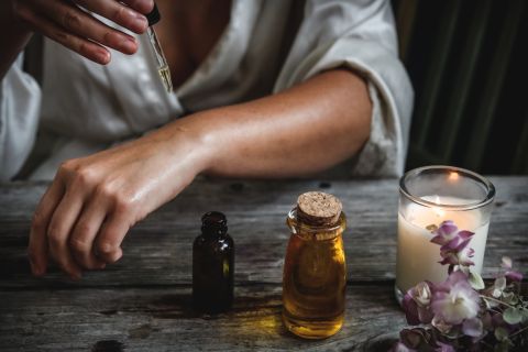 Eine Person in einer Robe trägt mit einer Pipette Öl auf ihren Arm auf. Die Szene umfasst eine kleine Glasflasche mit einem Korken, eine weitere Flasche, eine brennende Kerze und einige violette Blumen auf einem Holztisch.