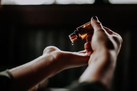 Ein Paar Hände hält eine kleine Bernsteinflasche, in deren Handfläche ein Tropfen Flüssigkeit fällt. Der Hintergrund ist sanft verschwommen, was eine beruhigende und intime Atmosphäre schafft.