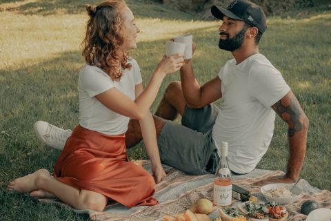 Ein Paar sitzt auf einer Picknickdecke und stößt mit Tassen an. Sie sind umgeben von einer Auswahl an Obst, Käse und einer Flasche Wein. Sie schauen sich an und lächeln, während sie einen sonnigen Tag im Freien genießen.