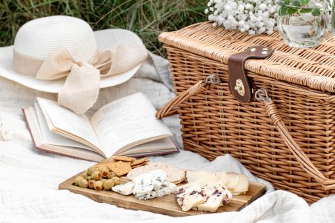 Ein gemütliches Picknick-Set besteht aus einem Weidenkorb, einem offenen Buch, einem Strohhut mit Schleife und einem Holzbrett mit verschiedenen Käsesorten, Crackern und Oliven, alles auf einer weißen Decke. Eine kleine Vase mit Blumen rundet die Szene ab.