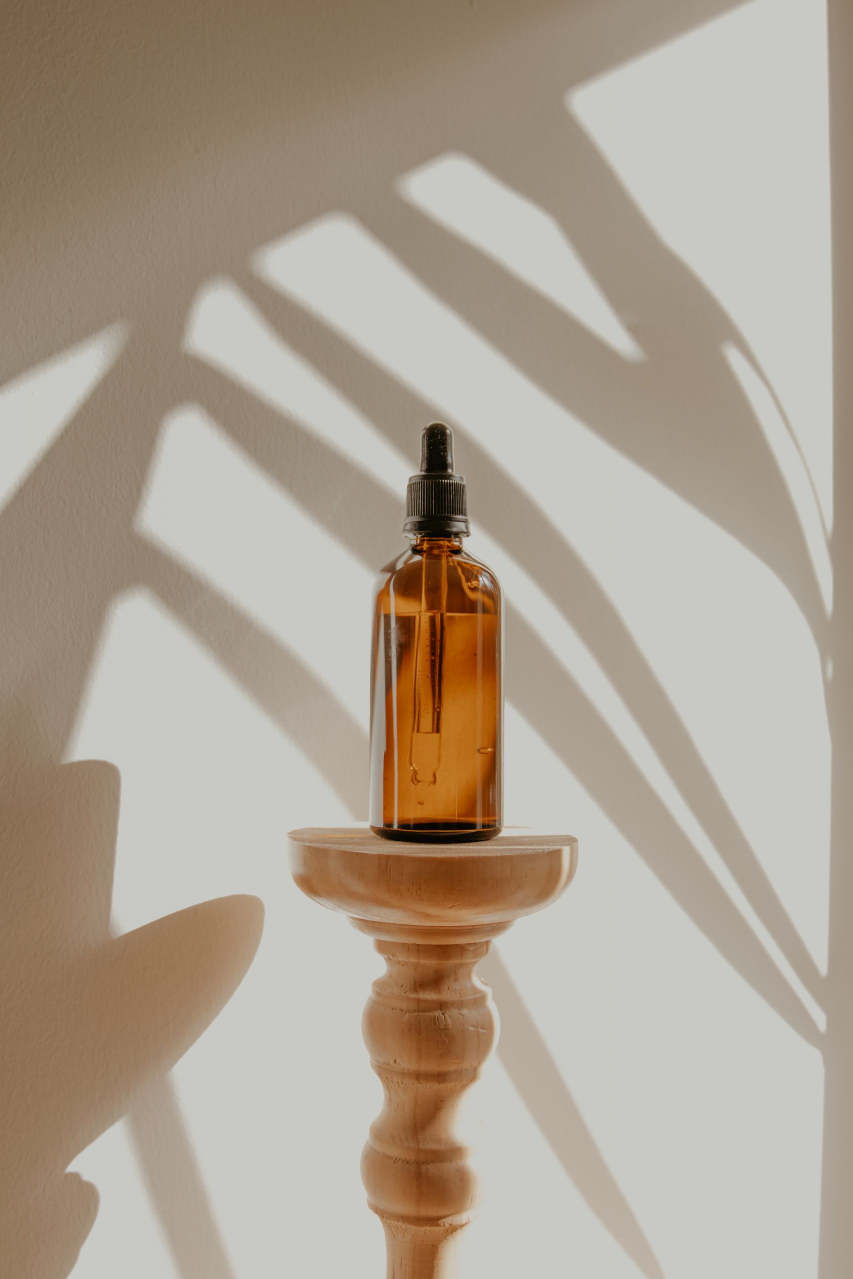Eine braune Glasflasche mit Pipette steht auf einem Holzständer vor einem hellen Hintergrund. Schatten von Blättern werden an die Wand geworfen und erzeugen einen natürlichen, künstlerischen Effekt.
