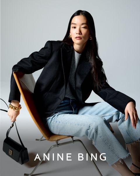 Eine Person sitzt in einem modernen Stuhl und trägt einen schwarzen Blazer, ein graues Hemd und hellblaue Jeans. Sie hält eine schwarze Handtasche mit einem goldenen Kettenriemen. Unten im Bild steht der Text „ANINE BING“.