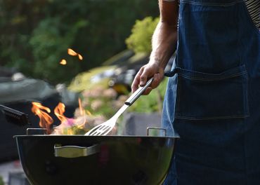 Eine Person mit einer Jeansschürze hält einen Spatel, während sie auf einem Holzkohlegrill mit Flammen grillt. Der Hintergrund ist mit grünem Laub verschwommen und die Szene vermittelt den Eindruck einer Kochsituation im Freien.
