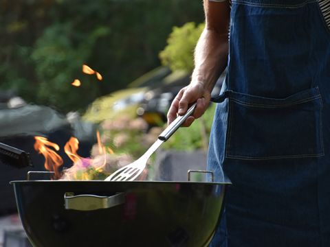 Eine Person mit einer Jeansschürze hält einen Spatel, während sie auf einem Holzkohlegrill mit Flammen grillt. Der Hintergrund ist mit grünem Laub verschwommen und die Szene vermittelt den Eindruck einer Kochsituation im Freien.