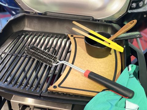 Eine Nahaufnahme eines Grills mit einem hölzernen Schneidebrett darauf. Auf dem Grill liegen eine Reinigungsbürste, eine Zange, ein schwarzer Topf und ein grünes Tuch. Die Szene suggeriert Vorbereitungen zum Kochen oder Grillen.