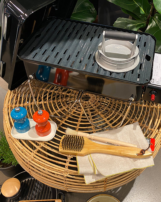 Ein Grill mit einem Brenner auf einem Korbtisch, dazu eine Grillbürste aus Holz, ein Tuch und kleine blaue und orangefarbene Gewürzmühlen auf einem Tablett. Im Hintergrund sind Pflanzen zu sehen.
