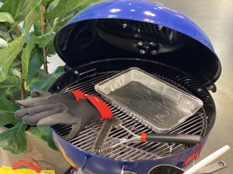 Ein blauer Holzkohlegrill mit offenem Deckel, auf dem eine Einweg-Tropfschale aus Aluminium zu sehen ist. Auf dem Grill liegen ein Paar grau-schwarze Handschuhe, eine Grillbürste und ein Spatel mit rotem Griff. Im Hintergrund sind grüne Blattpflanzen zu sehen.