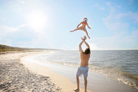 Ein Mann am Strand wirft ein fröhliches Baby in Ufernähe in die Luft. Die Sonne scheint hell am klaren blauen Himmel und wirft Spiegelbilder auf das ruhige Meer.