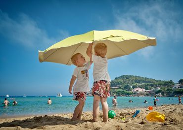 Zwei Kleinkinder in passenden Outfits stehen unter einem großen Sonnenschirm an einem sonnigen Strand. Im Hintergrund sind das Meer und mehrere schwimmende Menschen zu sehen, zusammen mit grünen Hügeln und einem klaren blauen Himmel.