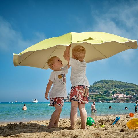 Zwei kleine Kinder in Shorts und T-Shirts halten einen großen gelben Sonnenschirm an einem Sandstrand. Im Hintergrund sind das Meer und schwimmende Menschen zu sehen, zusammen mit einem grünen Hügel und Häusern unter einem klaren blauen Himmel. In der Nähe liegen verstreut Strandspielzeuge.
