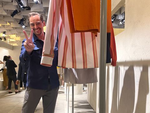 Ein lächelnder Mann, der ein Peace-Zeichen macht, steht neben einer Ausstellung gestreifter und einfarbiger Kleidung in einem modernen, gut beleuchteten Ausstellungsraum. Im Hintergrund sind weitere Personen zu sehen, die mit der Ausstellung interagieren.