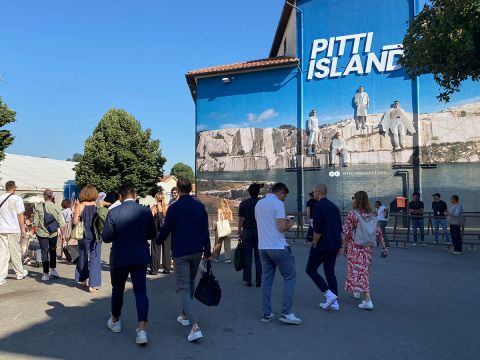 Eine Gruppe von Menschen geht draußen vor einem großen blauen Gebäude spazieren, auf dem „PITTI ISLAND“ steht. Auf dem Gebäude ist ein Foto von Männern in weißer Kleidung zu sehen. Im Hintergrund sind Bäume und ein klarer blauer Himmel zu sehen.