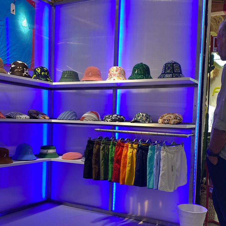 Eine Person steht vor einer Vitrine, in der auf den oberen Regalen verschiedene bunte Hüte ausgestellt sind und darunter bunte Shorts hängen. Die Vitrine wird von blauem Licht beleuchtet, das die Szene in ein leuchtendes Licht taucht.