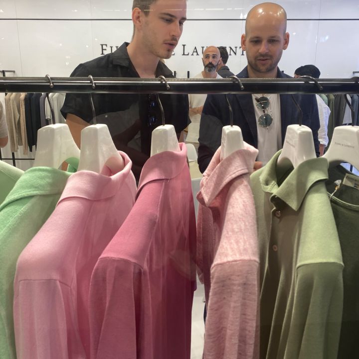 Zwei Personen schauen sich in einem Bekleidungsgeschäft Regale mit bunten Poloshirts an, darunter rosa, hellrosa, grün und hellgrün, die auf weißen Kleiderbügeln hängen. Andere stöbern in der Nähe. Im Hintergrund ist eine weiße Wand mit Text zu sehen.