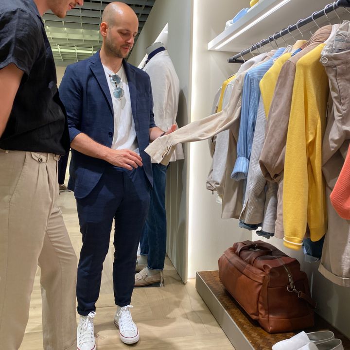 Zwei Männer stehen in einem Bekleidungsgeschäft und stöbern in einem Regal mit bunter Kleidung. Ein Mann im blauen Anzug zeigt auf ein beiges Hemd, während der andere im schwarzen Hemd zusieht. Auf einem Regal darunter sind eine braune Ledertasche und weiße Schuhe zu sehen.