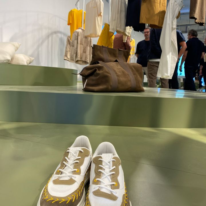 Im Vordergrund ist ein Paar stylische Sneaker mit weißen und gelben Akzenten zu sehen. Im Hintergrund sind auf Plattformen Kleidungsstücke in neutralen Tönen und eine braune Tasche ausgestellt, die von Menschen beobachtet werden.