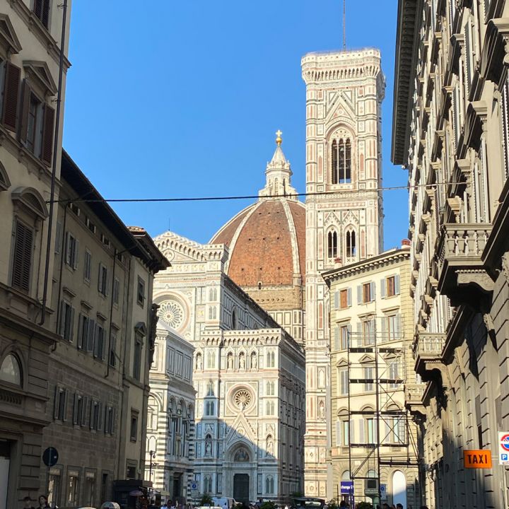 Straßenansicht von Florenz, Italien, mit der Kathedrale von Florenz, deren ikonische Kuppel und Glockenturm hervorstechen. Die Szene ist lebhaft mit Passanten und Autos, die entlang der schmalen Straße parken. Der Himmel ist klar und blau.