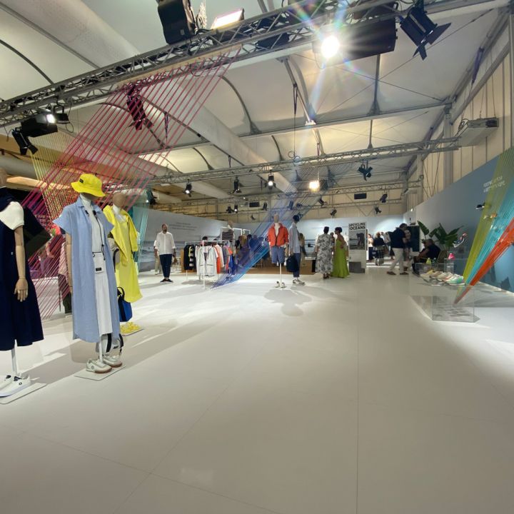 Ein geräumiger Modeschauraum mit Schaufensterpuppen in farbenfrohen, modernen Outfits. Helles Licht und eine hohe Decke schaffen eine offene Atmosphäre, während im Hintergrund Menschen in ausgestellten Kleidern stöbern.