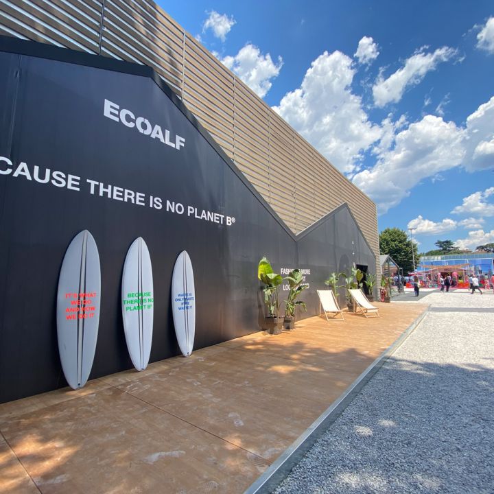Außenszene bei einer Ecoalf-Veranstaltung mit einer großen schwarzen Wand mit dem Slogan „Weil es keinen Planeten B gibt“ und Surfbrettern. Der Bereich verfügt über Liegestühle und Topfpflanzen unter einem blauen Himmel mit flauschigen Wolken.