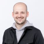 Vor einem schlichten weißen Hintergrund steht ein lächelnder Mann mit rasiertem Kopf, der eine schwarze Jacke über einem grauen Kapuzenpullover trägt.