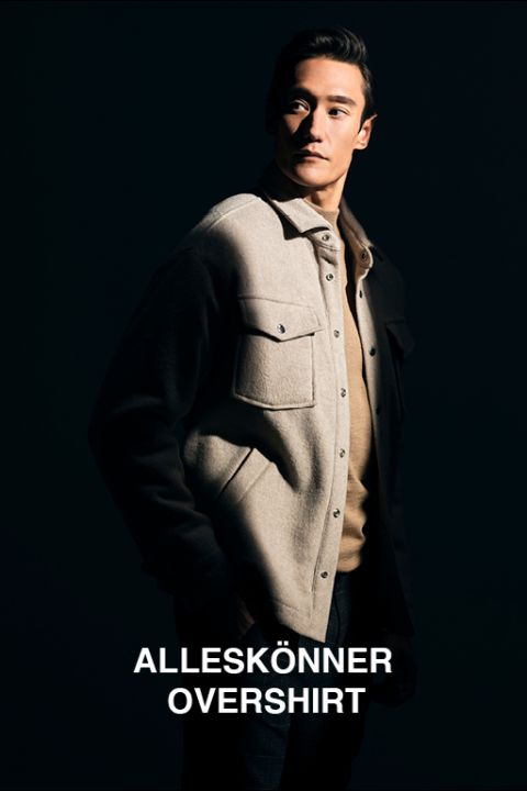 Eine Person steht selbstbewusst in einem schwach beleuchteten Raum und trägt ein helles Oberhemd über einem Pullover. Der Hintergrund ist dunkel, was einen dramatischen Kontrast erzeugt. Der Text unten lautet „ALLESKÖNNER OVERSHIRT“.