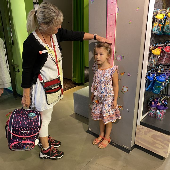 Eine Frau misst die Größe eines jungen Mädchens anhand einer Waage an einer Wand in einem Geschäft. Die Frau hält einen bunten Rucksack und das Mädchen steht aufrecht und trägt ein geblümtes Kleid. In der Nähe sind auf Regalen Wasserflaschen und Kleidung ausgestellt.