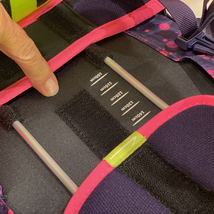 Nahaufnahme der Hand einer Person, die das Gurtsystem eines Rucksacks einstellt. Im Inneren befinden sich Klettverschlussfächer mit unterschiedlichen Größenbezeichnungen. Der Rucksack ist in den Farben Rosa und Lila mit einem gepunkteten Muster gestaltet.
