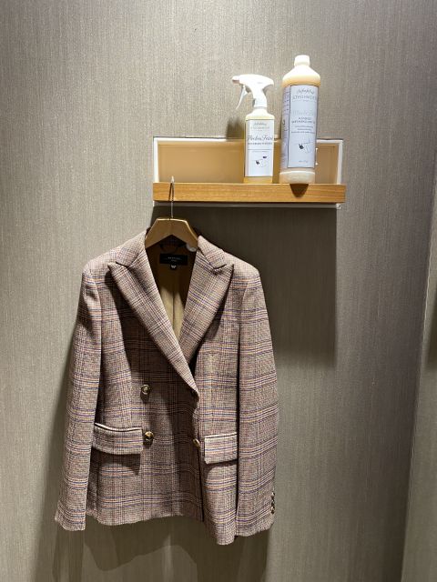 Ein karierter Blazer hängt auf einem hölzernen Kleiderbügel an einer strukturierten Wand. Darüber befindet sich ein kleines Regal mit einer Sprühflasche und einer Reinigungslösung.