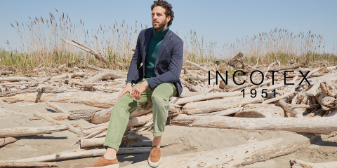 Ein Mann sitzt auf Treibholz an einem Strand. Er trägt einen marineblauen Blazer, einen grünen Pullover und eine grüne Hose. Dazu trägt er braune und weiße Schuhe. Im Hintergrund sind hohes Gras und ein klarer blauer Himmel zu sehen. Rechts steht der Text „INCOTEX 1951“.