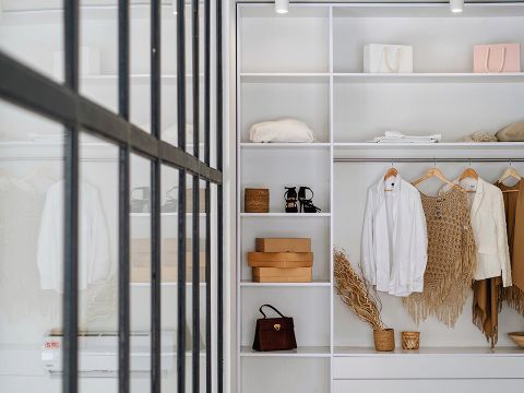 Ein ordentlich organisierter Kleiderschrank mit weißen Regalen und Kleiderbügeln. Er enthält Kleidung, Handtaschen und braune Kisten. Eine Glastrennwand spiegelt teilweise den Innenraum und trägt zum modernen Design bei.