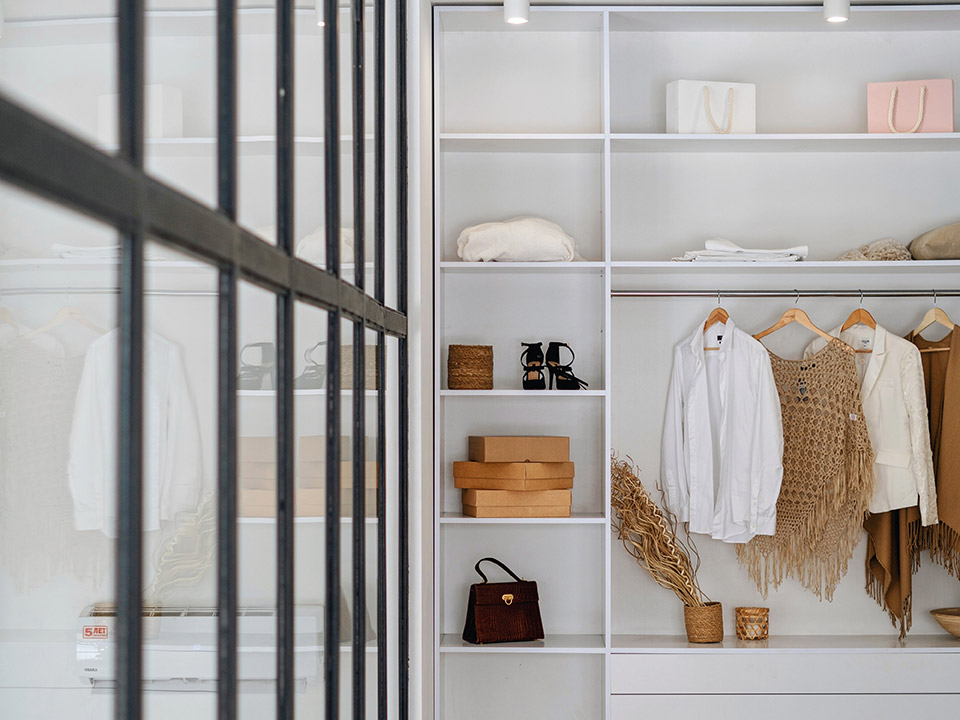 Ein ordentlich organisierter Kleiderschrank mit weißen Regalen und Kleiderbügeln. Er enthält Kleidung, Handtaschen und braune Kisten. Eine Glastrennwand spiegelt teilweise den Innenraum und trägt zum modernen Design bei.