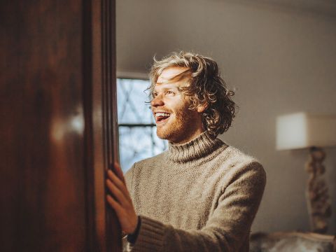 Eine Person mit lockigem Haar und Bart trägt einen kuscheligen, beigen Rollkragenpullover. Sie lächelt mit freudigem Gesichtsausdruck und schaut zur Seite. Der Raum ist warm beleuchtet, im Hintergrund sind ein Fenster und eine Lampe.