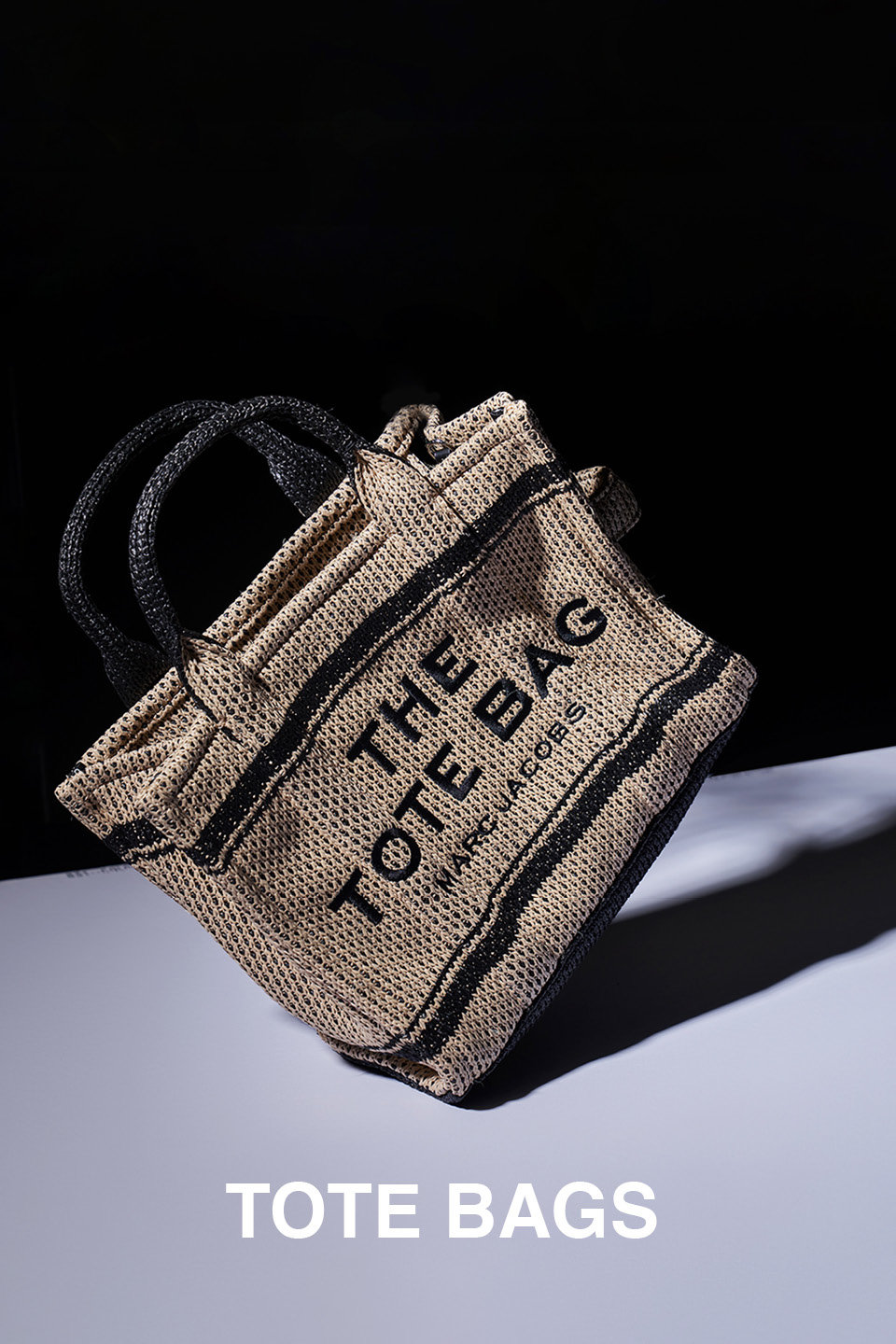 Eine gewebte Einkaufstasche, die bei dramatischer Beleuchtung auf einer weißen Oberfläche präsentiert wird und einen Schatten wirft. Die Tasche hat schwarze Griffe und ist mit dem Text „THE TOTE BAG“ in großen Buchstaben und „MARC JACOBS“ in kleineren Buchstaben verziert.