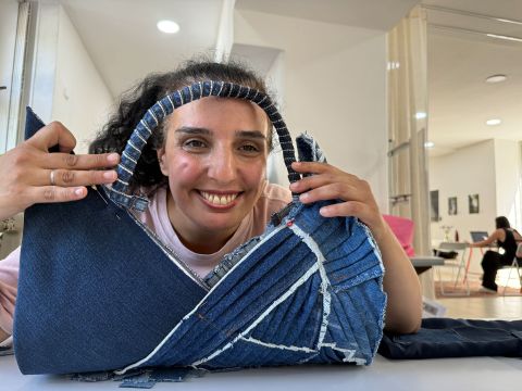 Eine lächelnde Frau sitzt an einem Tisch in einem hellen, modernen Raum und hält eine große, handgefertigte Jeans-Tasche mit Patchwork-Details in der Hand.