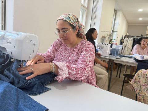 Eine Frau in einem rosa gemusterten Hemd näht mit einer Nähmaschine Jeansstoff, während andere an Tischen in einem hellen Raum arbeiten.