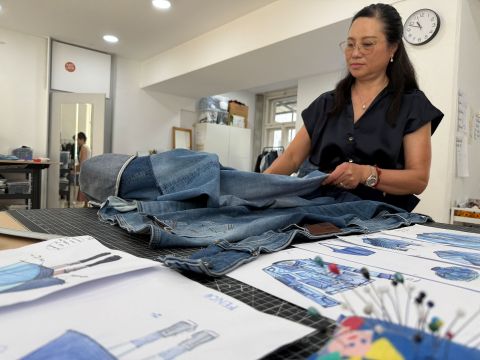 Eine Frau faltet Jeansstoff an einem Arbeitsplatz, an dem Skizzen, Nähutensilien und eine Uhr an der Wand zu sehen sind, in einem hellen, modernen Raum.