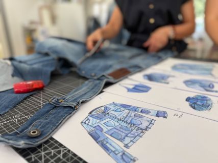Eine Person schneidet Jeansstoff zu, während auf dem Tisch daneben Modeskizzen von Patchwork-Jacken zu sehen sind.