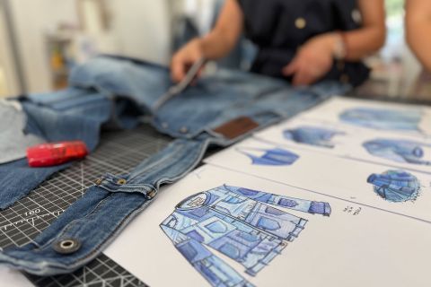 Eine Person schneidet Jeansstoff zu, während auf dem Tisch daneben Modeskizzen von Patchwork-Jacken zu sehen sind.
