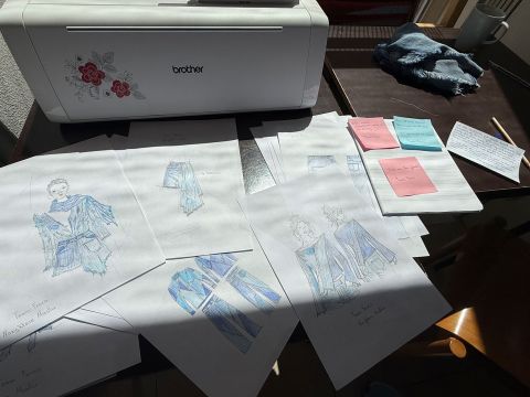 Auf einem Tisch neben einer Brother-Schneidemaschine liegen Nähskizzen und Notizen verstreut, daneben befinden sich Haftnotizen und Stoffmuster im Sonnenlicht.