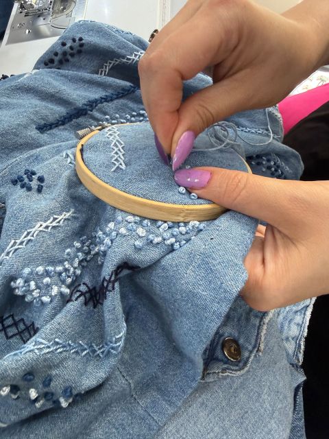 Hände mit lila Fingernägeln sticken mit einem Stickrahmen weiße und blaue Fäden auf Jeansstoff.