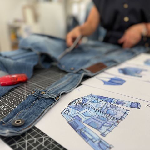 Eine Person schneidet neben einer farbenfrohen Jackenskizze auf einem Tisch Jeansstoff zu, daneben liegen Nähutensilien.