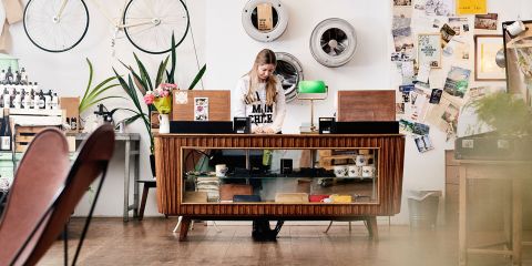 Eine Person steht hinter einer Vintage-Holztheke in einem Laden im Retro-Stil, umgeben von Fahrrädern, Wandkunst und Pflanzen. Der Laden ist vielseitig eingerichtet, darunter Fotos und ein an der weißen Wand montierter Metallventilator.
