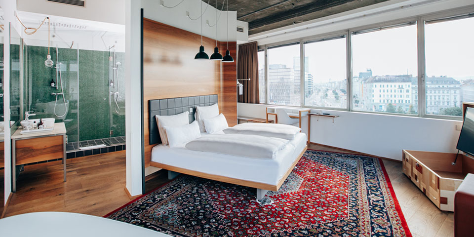 Geräumiges, modernes Hotelzimmer mit großem Bett auf gemustertem Teppich, Dusche mit Glaswänden und Blick auf die Stadt durch breite Fenster. Die zeitgenössische Einrichtung umfasst Hängelampen, Holzakzente und eine helle Farbpalette.