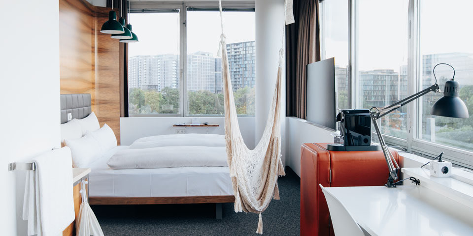 Ein modernes Hotelzimmer mit großen Fenstern verfügt über eine Hängematte, ein Bett mit weißer Bettwäsche, einen Schreibtisch mit Lampe und eine Kaffeemaschine. Auf einem Schrank steht ein Fernseher, und draußen sind städtische Gebäude zu sehen.