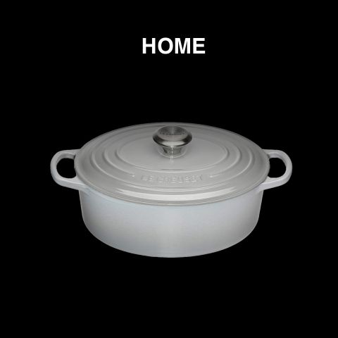 Ein weißer Le Creuset Schmortopf mit Deckel und zwei Griffen, zentriert auf schwarzem Hintergrund. Über dem Topf steht in weißer Schrift das Wort „HOME“.