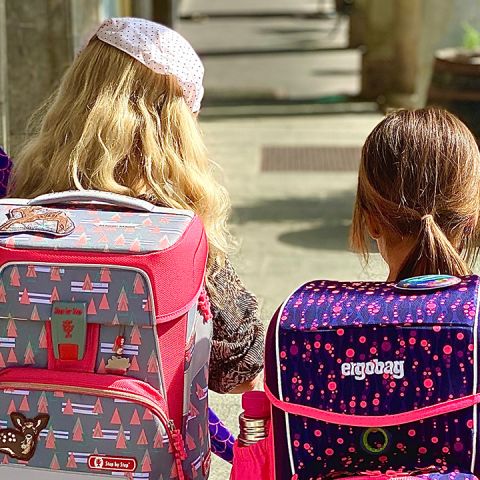 Zwei Kinder gehen mit bunten Rucksäcken spazieren. Das Kind links hat einen rot-braunen Rucksack mit Baummustern, das Kind rechts einen lila Rucksack mit kleinen rosa Formen. Beide tragen Freizeitkleidung und gehen von der Kamera weg.