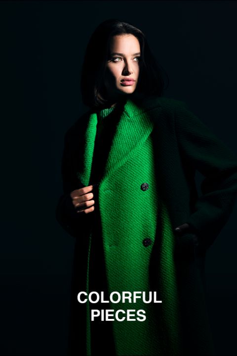 Eine Frau steht in gedämpftem Licht, trägt einen hellgrünen Mantel und blickt intensiv. Der Mantel hat einen übergroßen Kragen und ein zweireihiges Design. Unten steht der Text „Colorful Pieces“.